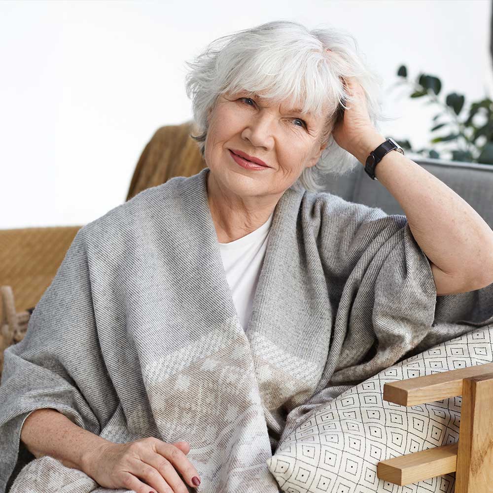 older woman wrapped in a grey ponch sitting in a comfy chair while looking thoughtful
